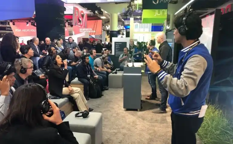 A group of people wearing headphones listen attentively to a presenter at a tech expo. The presenter gestures with his hands, while attendees sit on benches surrounded by booths and colorful displays.