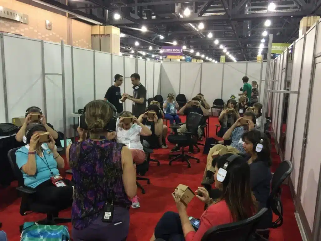 A group of people seated on chairs in a room with red carpet wear headphones and use virtual reality headsets. A presenter stands in front of them, guiding the immersive experience. Partitioned booths are visible in the background.