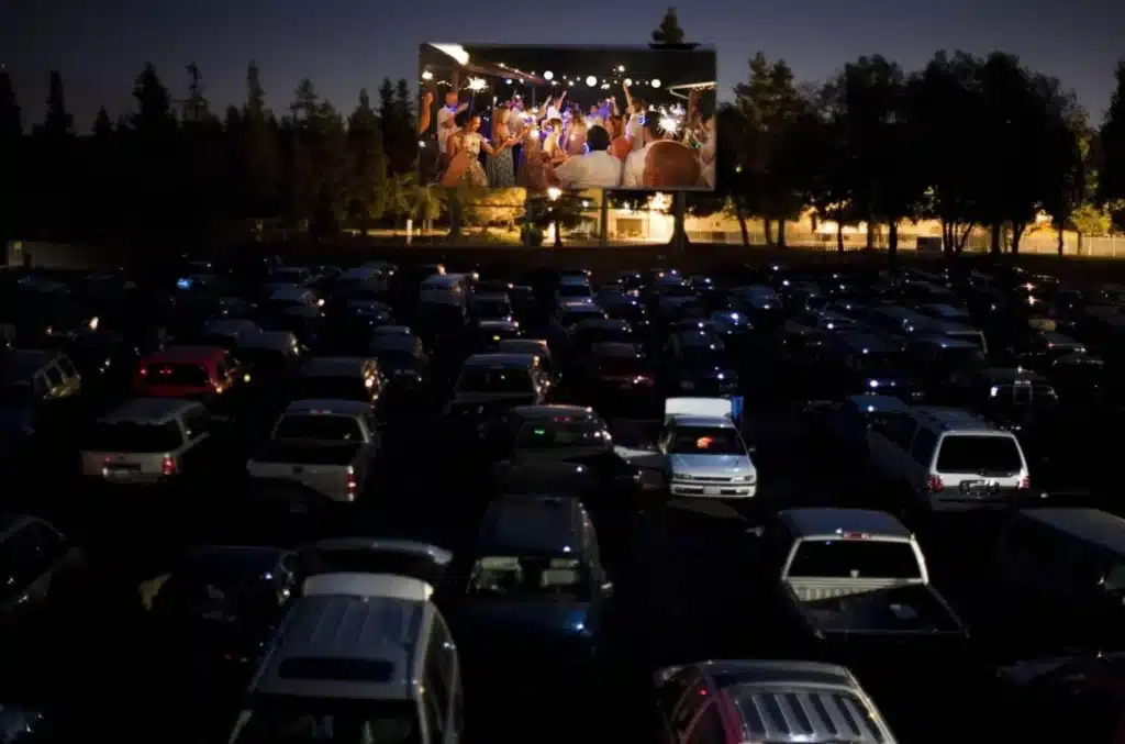 A nighttime scene at a drive-in theater shows many parked cars facing a large outdoor movie screen displaying a brightly lit party scene. Trees and a fence line the background.