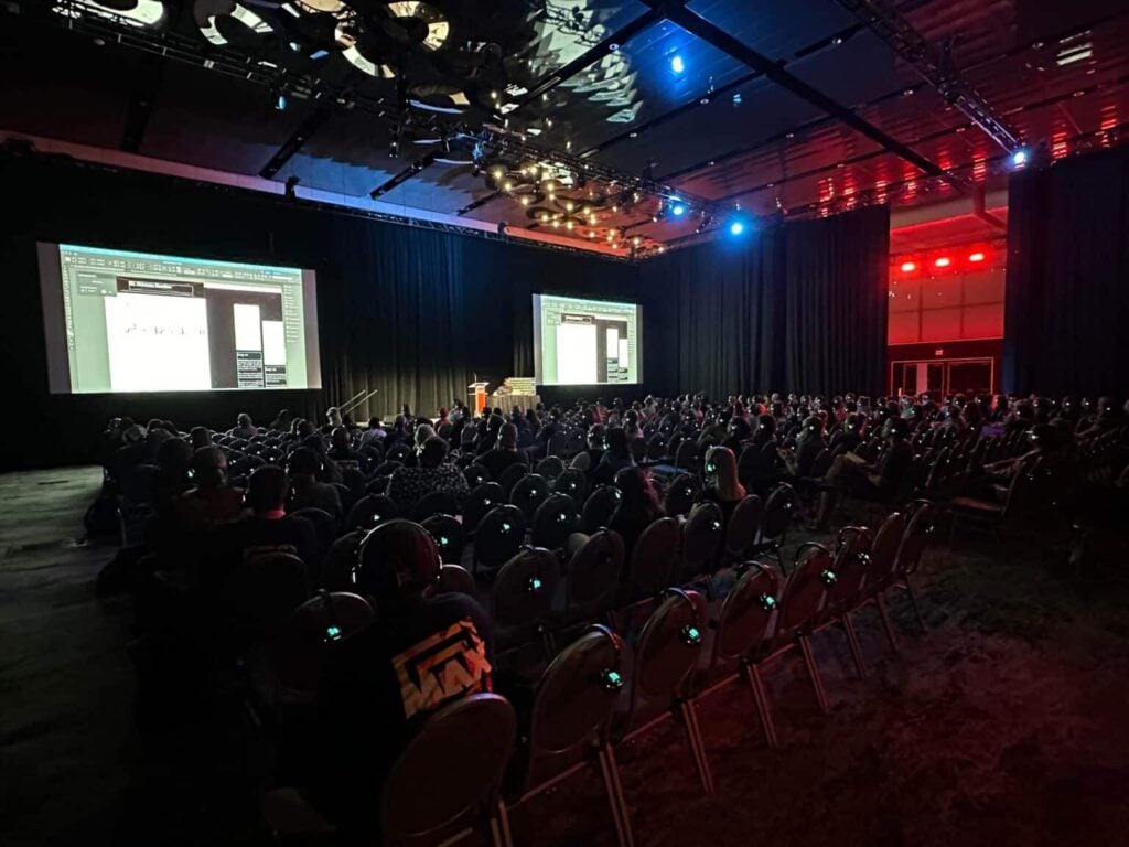 A large audience sits in a dark auditorium facing two projection screens and a speaker at a podium during a presentation, breaking language barriers with silent conferences and enhanced by red and blue lighting illuminating the room.